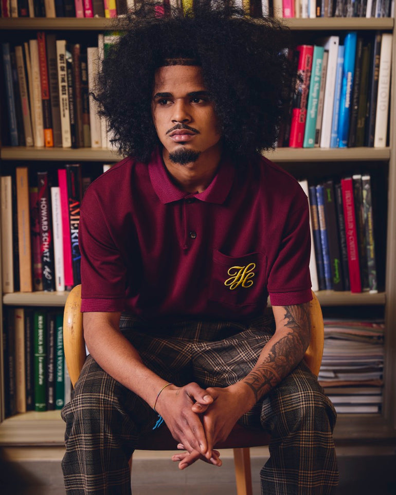 IH NOM UH NIT FW26 burgundy short-sleeve polo shirt with yellow embroidered monogram on chest pocket, front view.